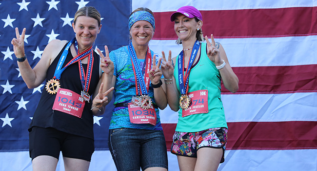 Three women posing together for a photo after receiving award medals in front of an American flag.